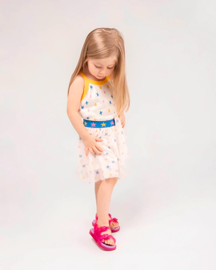 A young model posing for a picture in a white patterned dress and a pair of pink/lilac Mini Melissa Wide Sandal with a pink base, two pink buckles on top, a back ankle strap and a lilac insole.