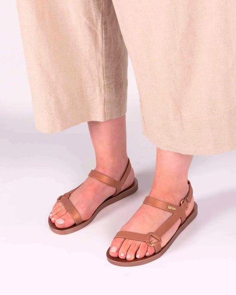 A model's legs in beige pants and a pair rose metallic Melissa Sun Downtown sandals with a front and ankle strap joined together by a vertical strap and a triangle buckle.