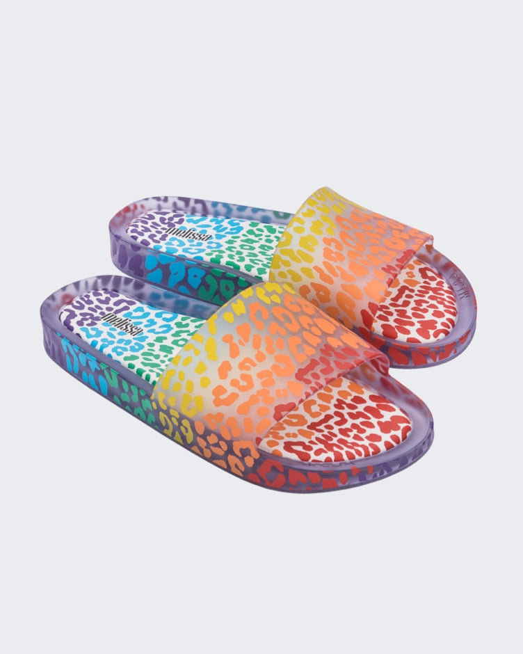 An angled top view of a pair of clear / orange Melissa Rainbow Beach slides with a clear base and a rainbow cheetah print pattern.