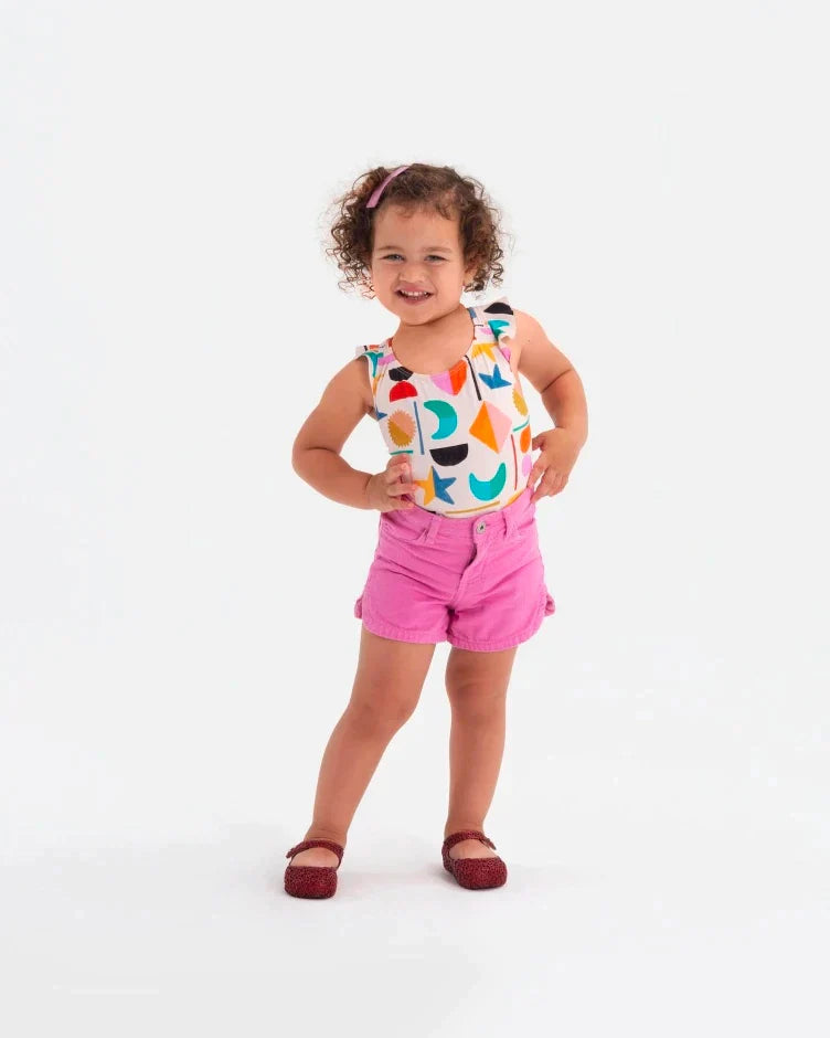 A child model posing for a picture in a multicolor patterened top, pink shorts and wearing a pair of red glitter Mini Melissa Campana flats with a snap strap for baby and an open woven texture.