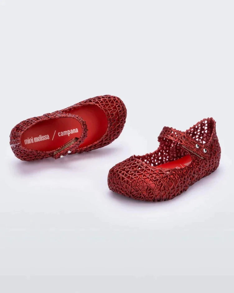 An angled side and top view of a pair of red glitter Mini Melissa Campana flats with a snap strap for baby and an open woven texture.