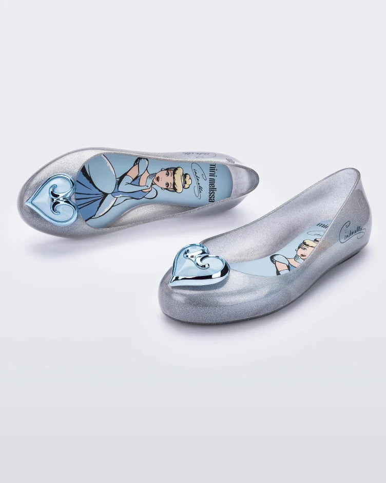 An angled side and top view of a pair of silver Mini Melissa Sweet Love Princess flats, with a silver base, Cinderella in script on the side, a heart detail on the toe, and a drawing of Cinderella on the sole.