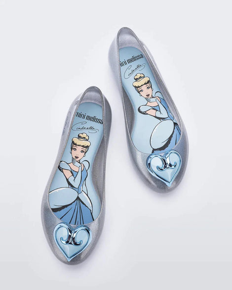 Top view of a pair of silver Mini Melissa Sweet Love Princess flats, with a silver base, Cinderella in script on the side, a heart detail on the toe, and a drawing of Cinderella on the sole.