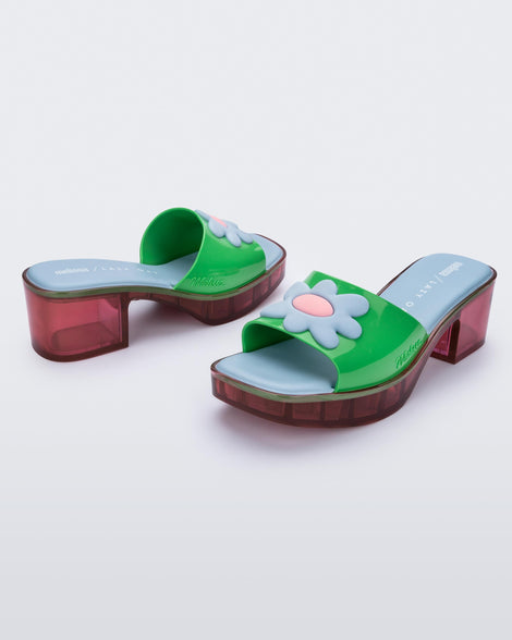 Angled view of a pair of multicolored Melissa Shape heeled slides, with transparent pink sole and green strap with blue and pink flower in the center.