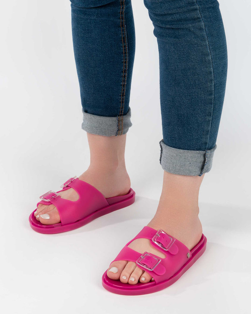 A model's legs in denim, wearing a pair of dark pink Melissa Wide Slides with two straps fastened at the top with two clear buckles.