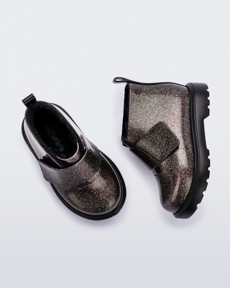 A side and top view of a pair of black/glitter multicolor Mini Melissa Chelsea boots with a multicolor glitter base, velcro front strap and a black sole.
