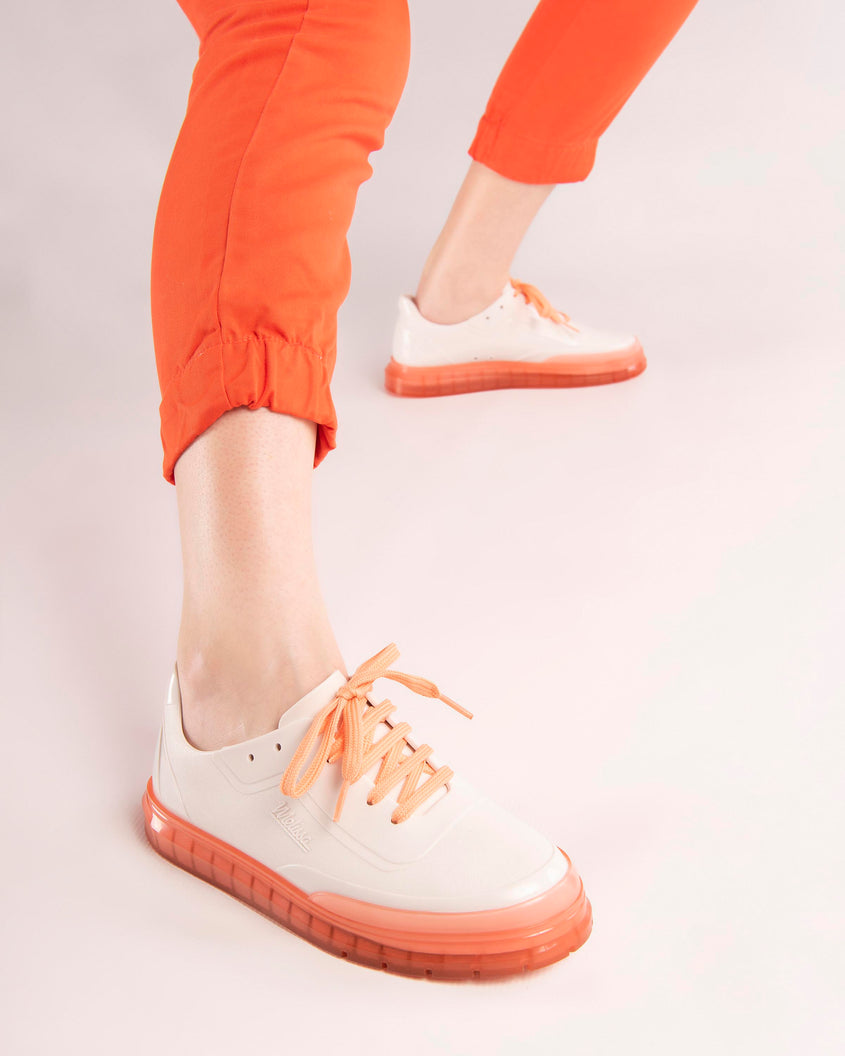 A model's legs wearing orange pants and a pair of white Melissa Classic sneakers with a white base, laces, orange sole and a melissa logo on the side.