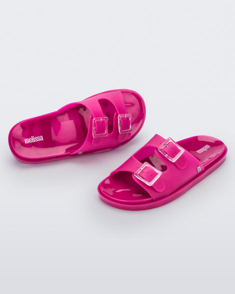 An angled front and top view of a pair of dark pink Melissa Wide Slides with two straps fastened at the top with two clear buckles.