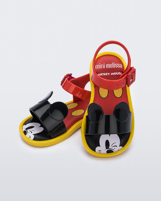 An angled top view of a red Mini Melissa Mar Sandal with mickey mouse insole, a red top strap, black bottom strap with mickey mouse ears on the top and a yellow sole, leaning on eachother.