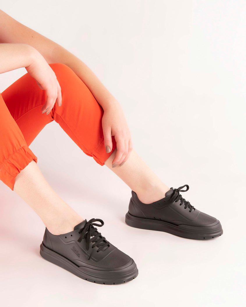 A model's legs wearing orange pants and a pair of matte black Melissa Classic sneaker with a black base, laces, sole and a melissa logo on the side.
