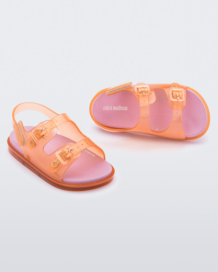 An angled front and top view of a pair of Orange/Pink Mini Melissa Wide Sandals with a clear orange base, two buckles at the top, a velcro ankle strap and a pink insole.