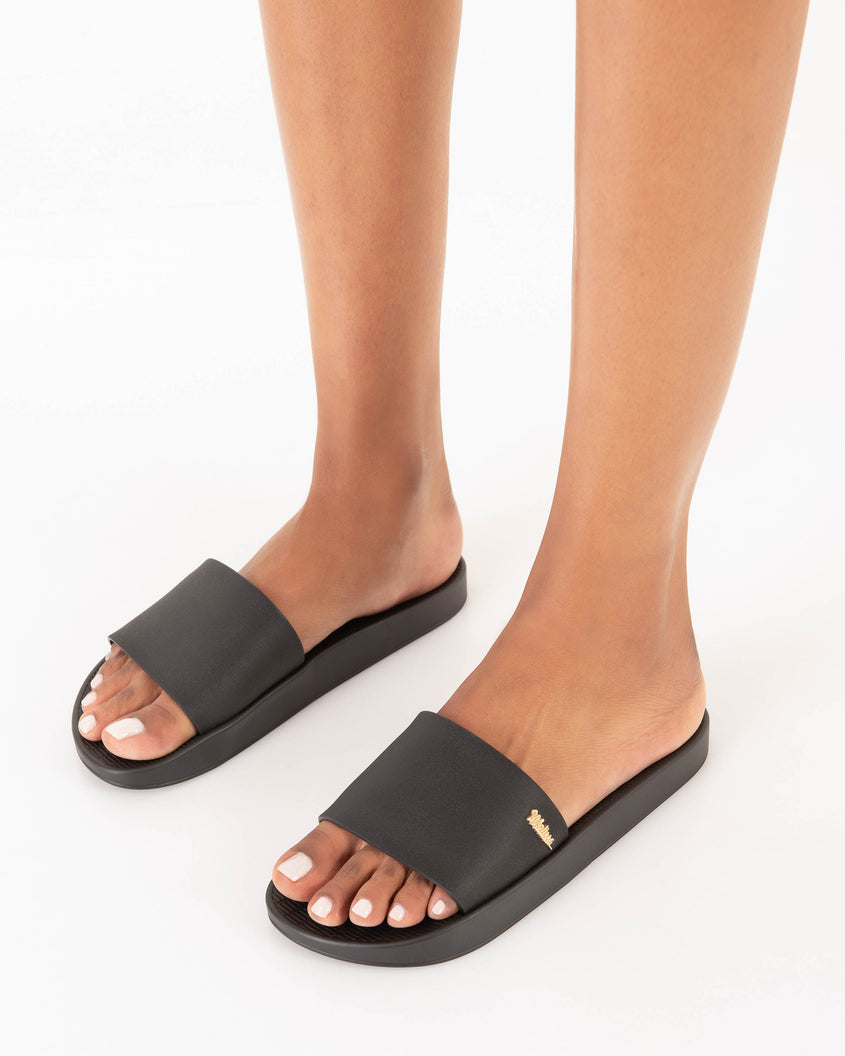 A model's legs wearing a pair of Melissa Sun Sunset slide sandals in black with gold Melissa logo on the top side corner of the strap. 