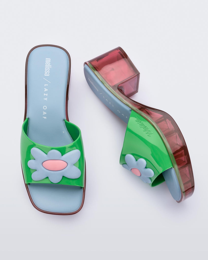 Top and side view of a pair of multicolored Melissa Shape heeled slides, with transparent pink sole and green strap with blue and pink flower in the center.