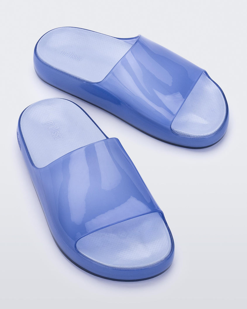 An angled front and top view of a pair of transparent blue Melissa Cloud slides.