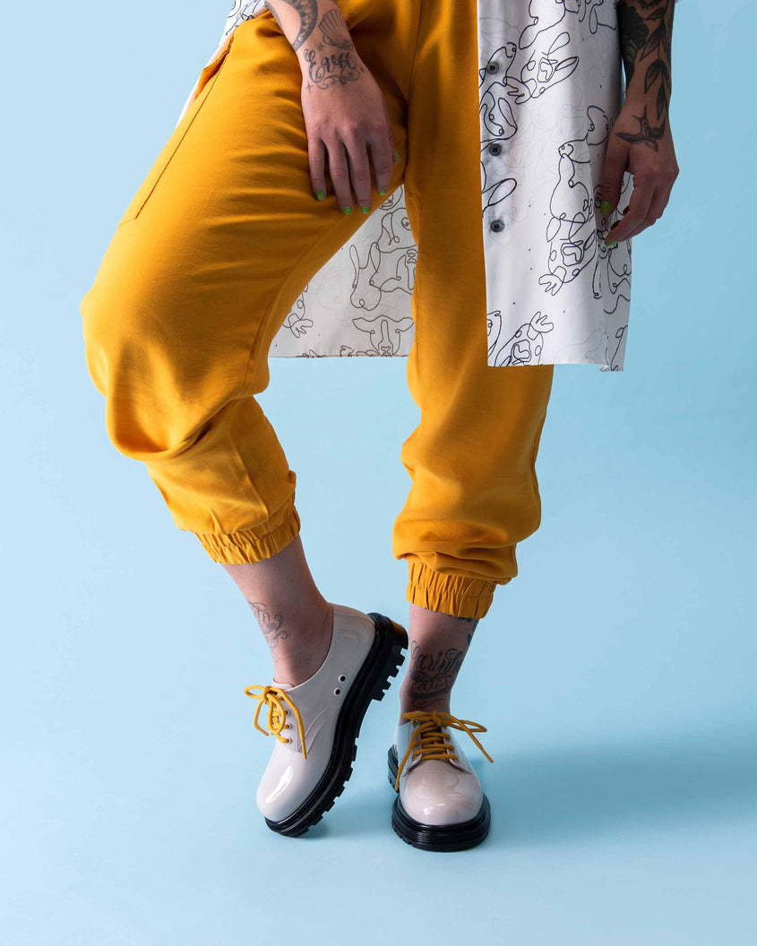 A model's legs in yellow pants and a white patterend top, wearing a pair of beige Melissa Bass sneakers with a beige base, yellow laces and a black sole.