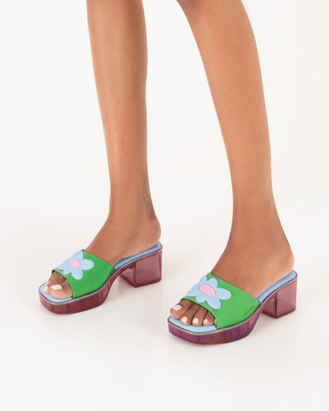 Model's legs wearing a pair of multicolored Melissa Shape heeled slides, with transparent pink sole and green strap with blue and pink flower in the center.