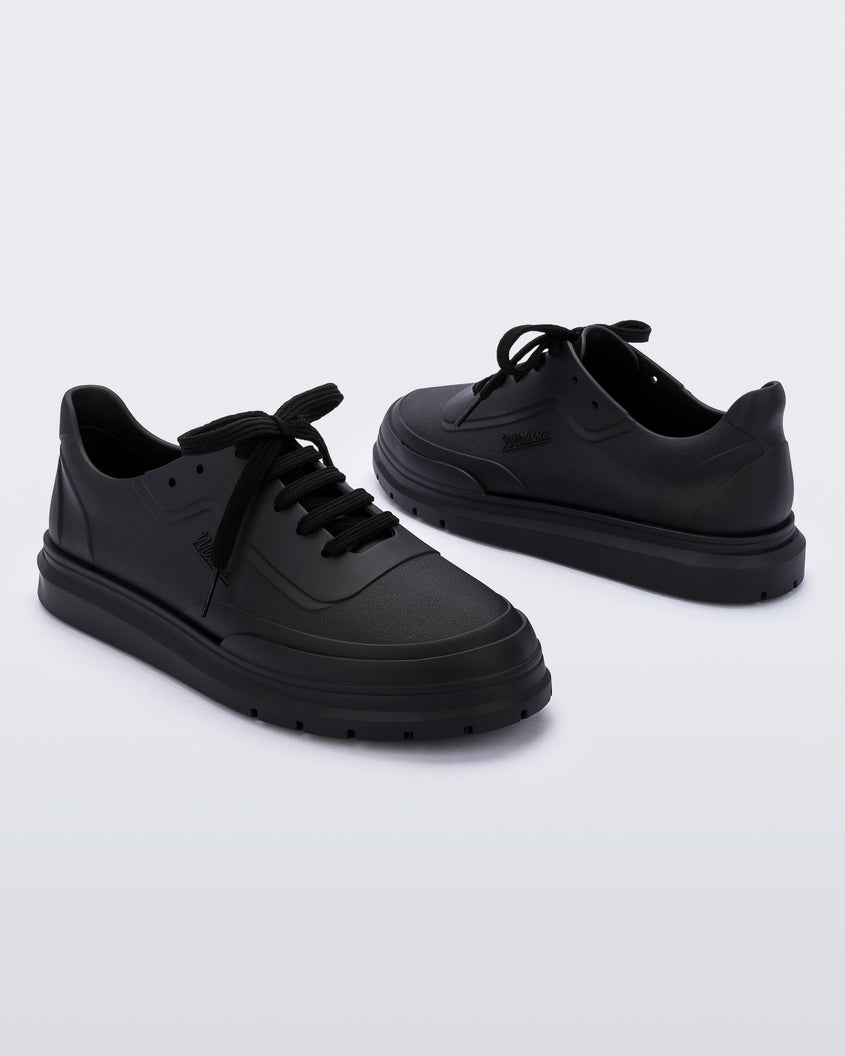 An angled inner and outter side view of a pair of matte black Melissa Classic sneakers with a black base, laces, sole and a melissa logo on the side.