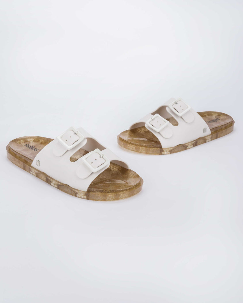 Angled view of a pair of Melissa Wide slide sandals with gold glitter sole and white double straps with buckles