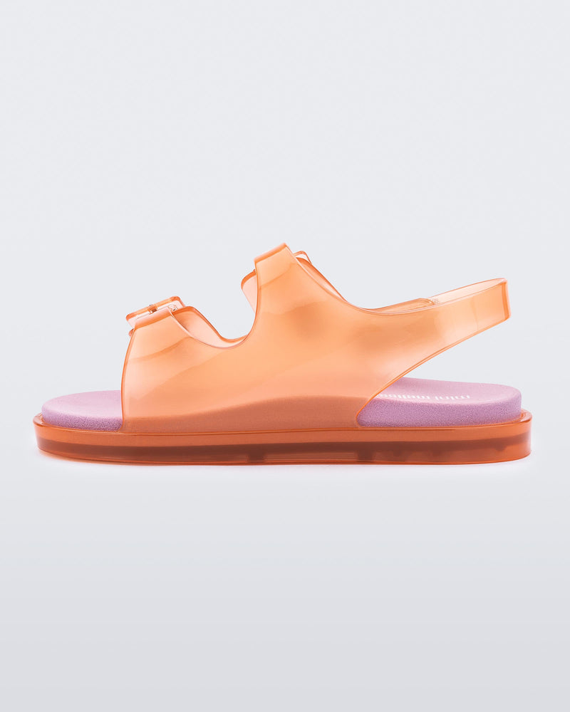 An inner side view of a Orange/Pink Mini Melissa Wide Sandal with a clear orange base, two buckles at the top, a velcro ankle strap and a pink insole.