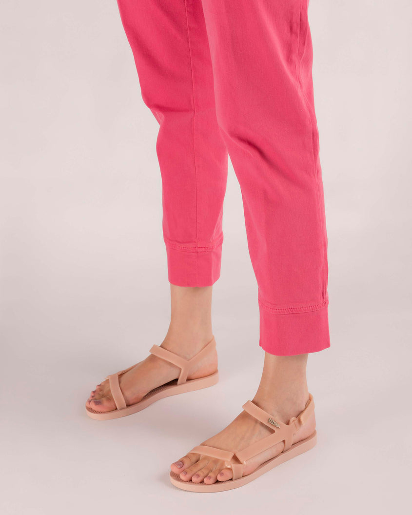 A model's legs in bright pink pants wearing a pair of Melissa Sun Downtown sandals with pink front cross and back ankle straps and a pink sole. 