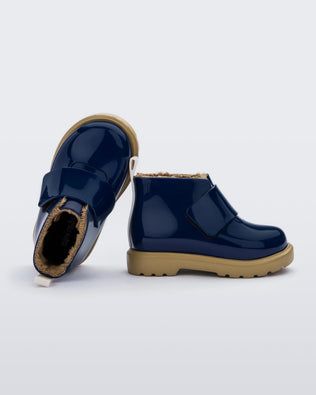 A top and side view, with one boot laying on it's back, of pair of blue/beige Mini Melissa Chelsea blue boots with a velcro strap, dark blue base, beige sole and beige fuzzy lining.