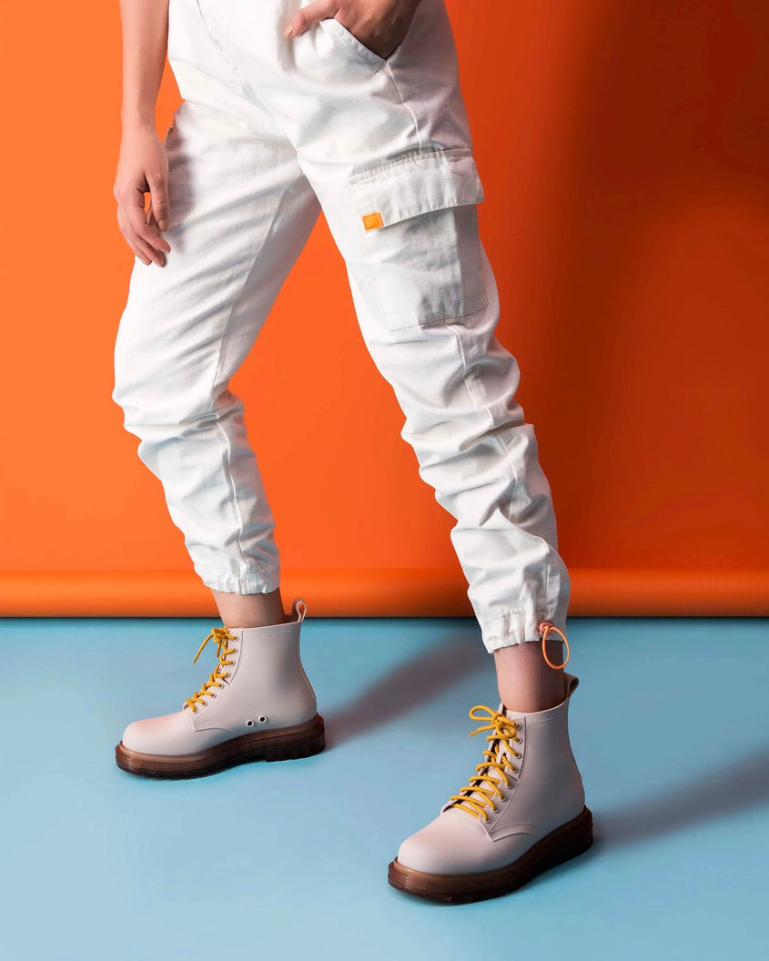A model's legs wearing white pants and a pair of matte beige/yellow Melissa Coturno boots, with a matte beige base, yellow laces and a brown sole, in front of an orange backdrop.