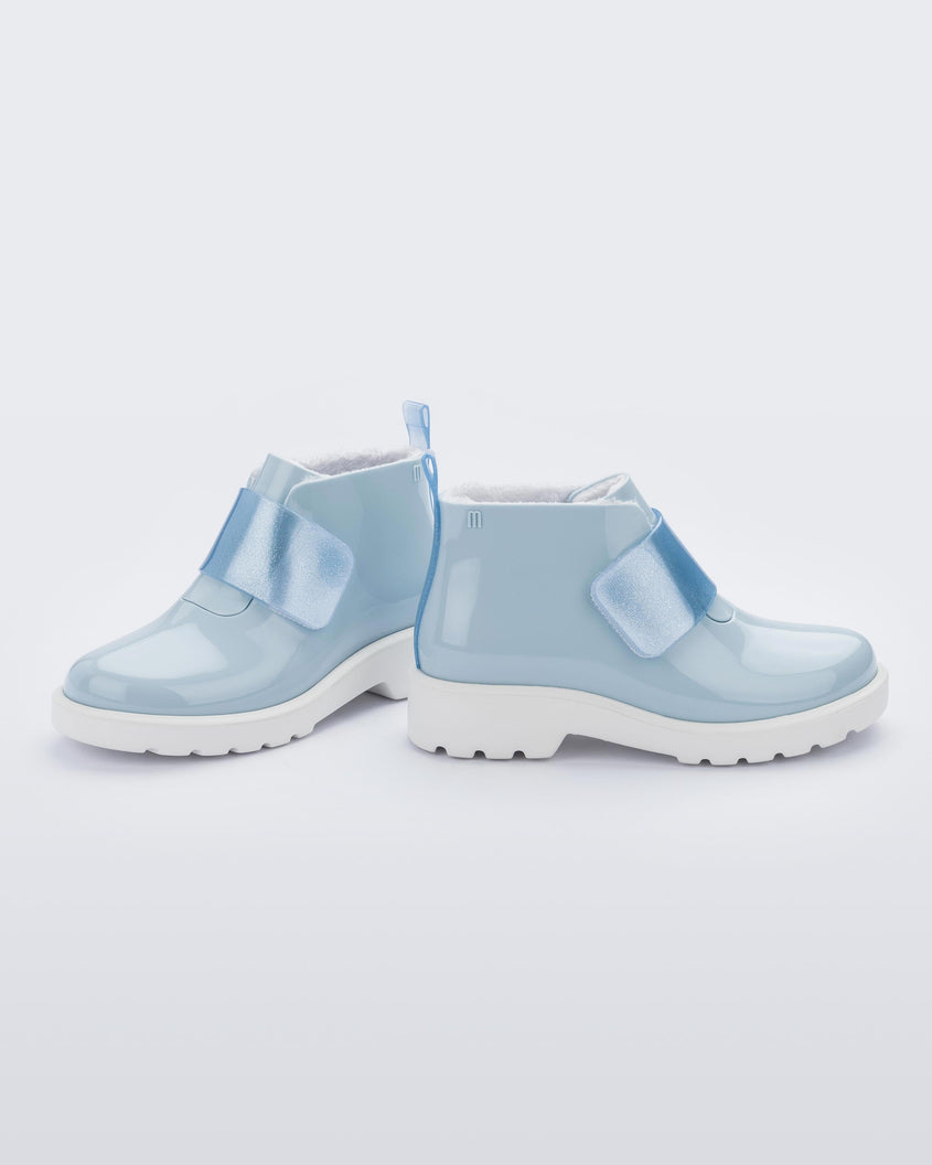 Side view of a pair of blue Mini Melissa Chelsea boots with a light blue base, glitter blue velcro front strap and a white sole.
