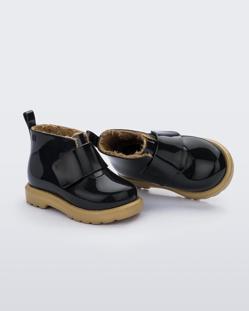 An angled top and side view of a pair of black/beige Mini Melissa Chelsea boots with a black base, velcro front strap and beige sole.