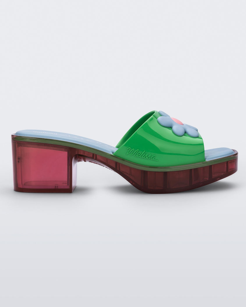 Side view of a multicolored Melissa Shape heeled slide, with transparent pink sole and green strap with blue and pink flower in the center.