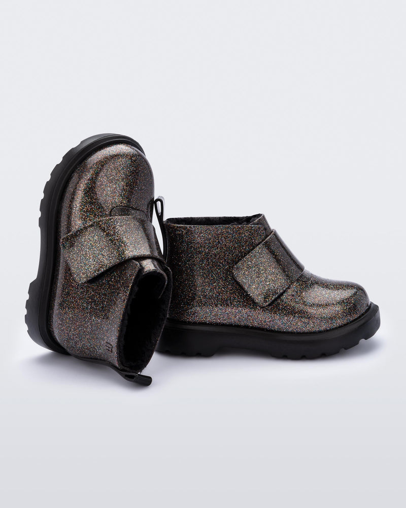 An angled side and top view of a pair of black/glitter multicolor Mini Melissa Chelsea boots with a multicolor glitter base, velcro front strap and a black sole.