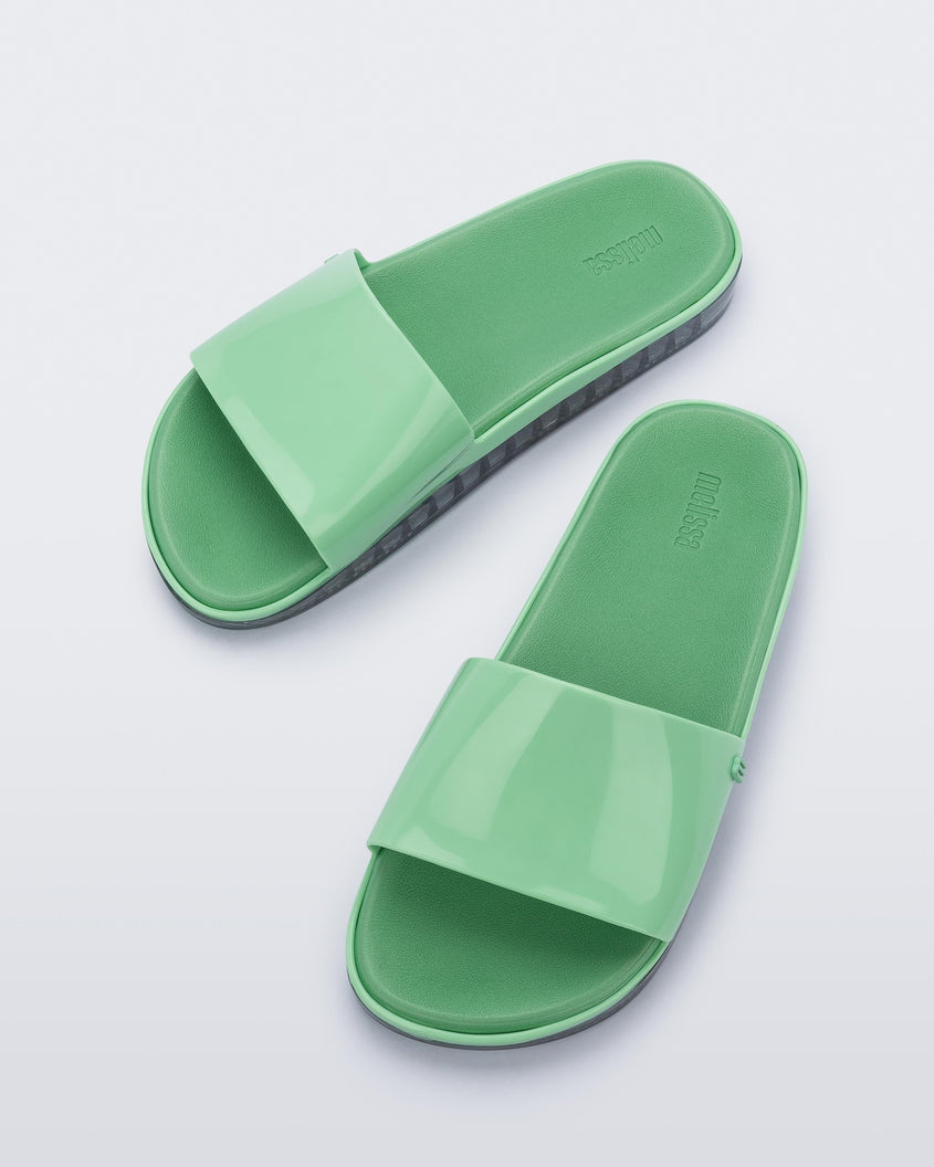 Angled view of a pair of green Melissa Beach slides with clear soles. 