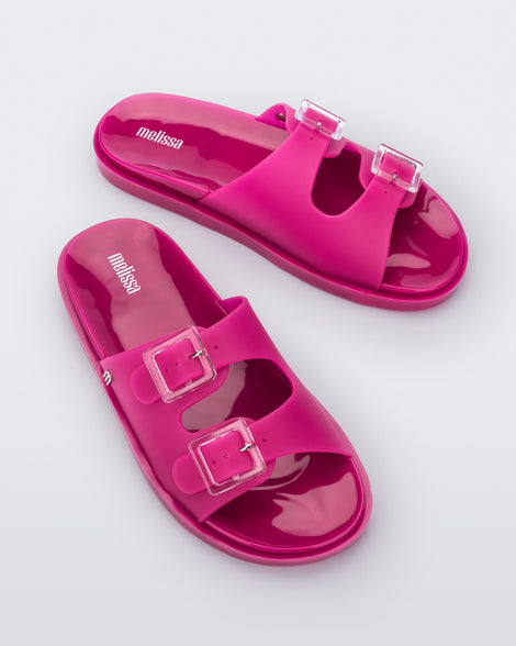 An angled top view of a pair of dark pink Melissa Wide Slides with two straps fastened at the top with two clear buckles.