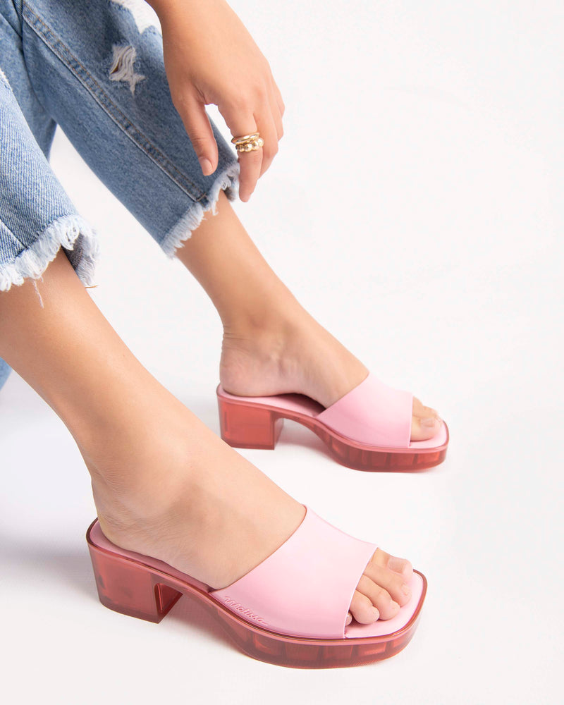 A model's legs in jeans wearing a pair of Melissa Shape slides with transparent pink platform heel and a pink wide front strap. 
