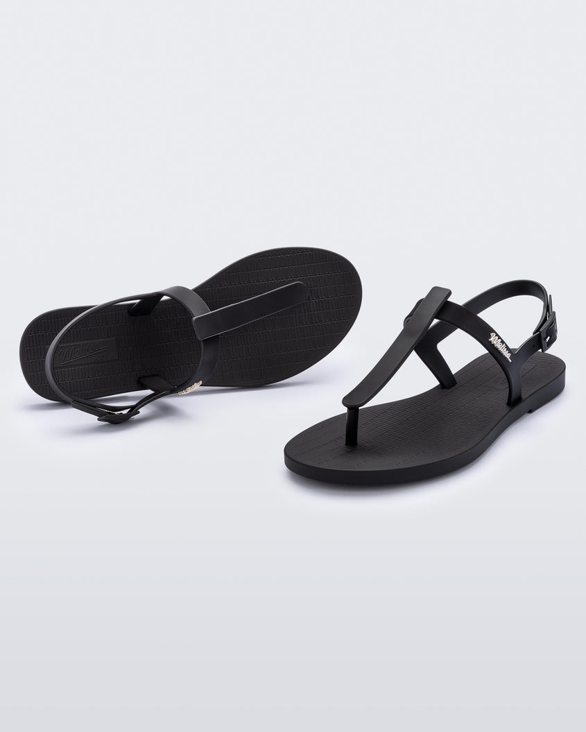 An angled front and top view of a pair of black Melissa Sun Ventura sandal with a top strap, reading melissa in silver, intersected by a vertical front strap detail and an ankle strap.