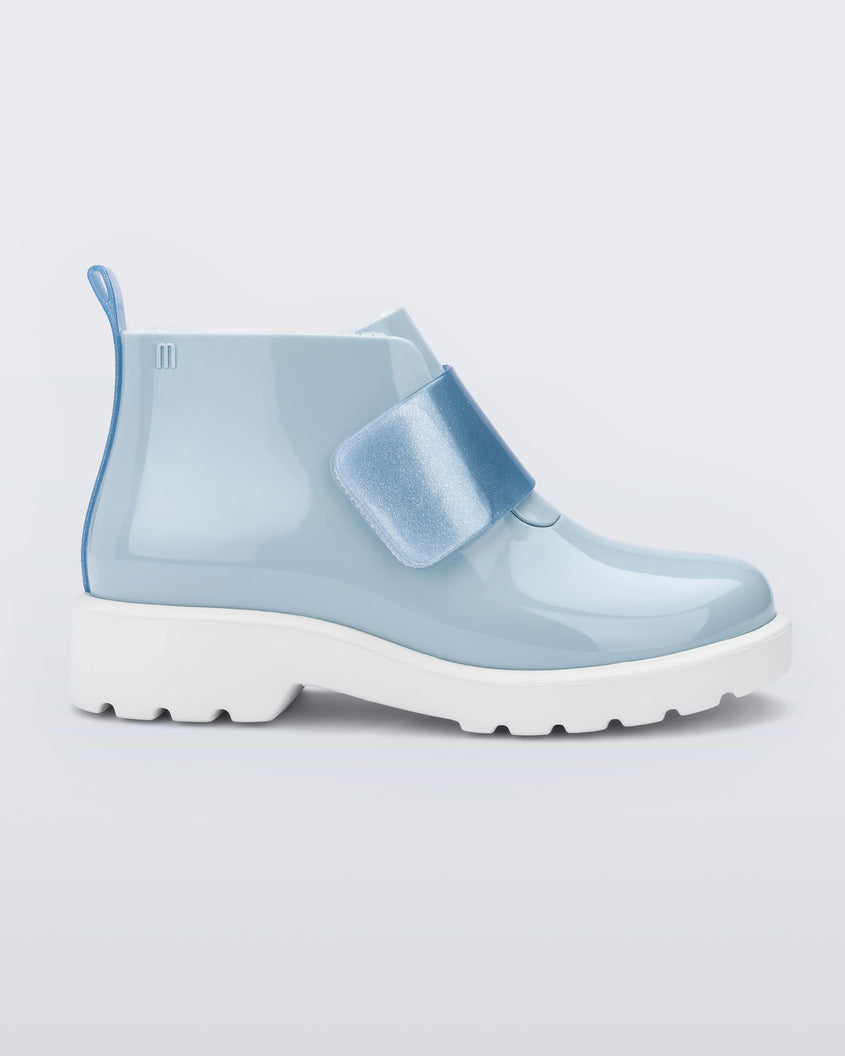 An outter side view of a blue Mini Melissa Chelsea boot with a light blue base, glitter blue velcro front strap and a white sole.