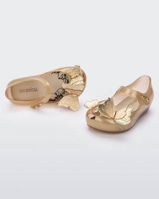 An angled front and top view of a pair of gold Mini Melissa Ultragirl Butterfly flats with a top strap and two butterfly details on the toe.