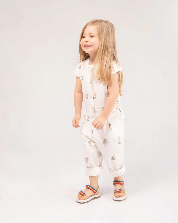 A little girl standing in a white outfit and wearing white Mini Melissa Papete sandals with rainbow straps.